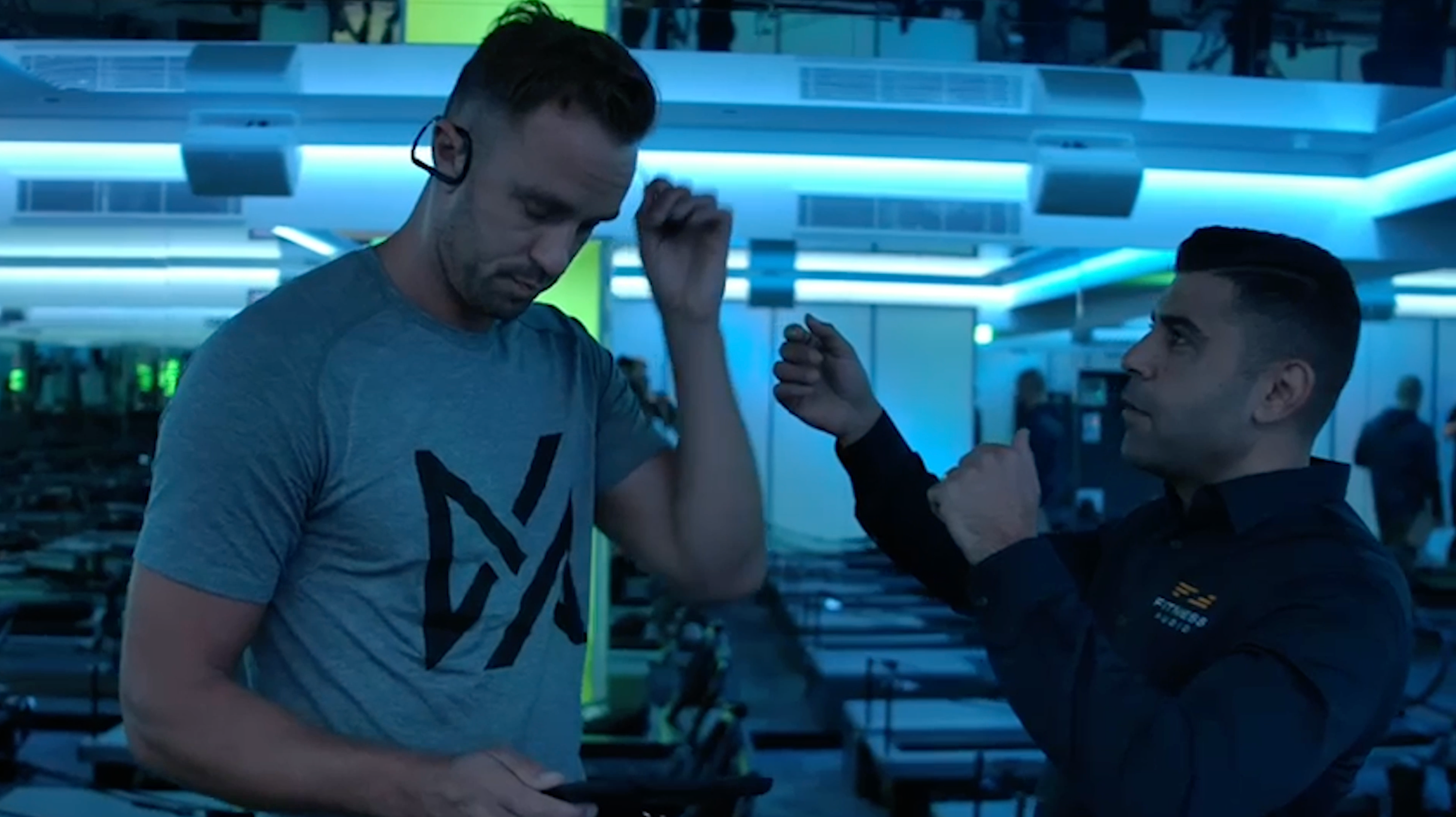 Two men in a gym setting, one wearing a blue t-shirt with a logo and the other in a dark jacket.