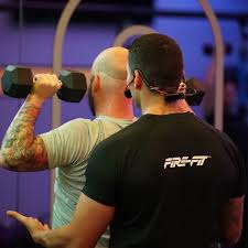 Fitness instructor coaching a gym goer lifting weights, with their backs to viewer displaying a red EMic and MTU8 transmitter on the instructors head.