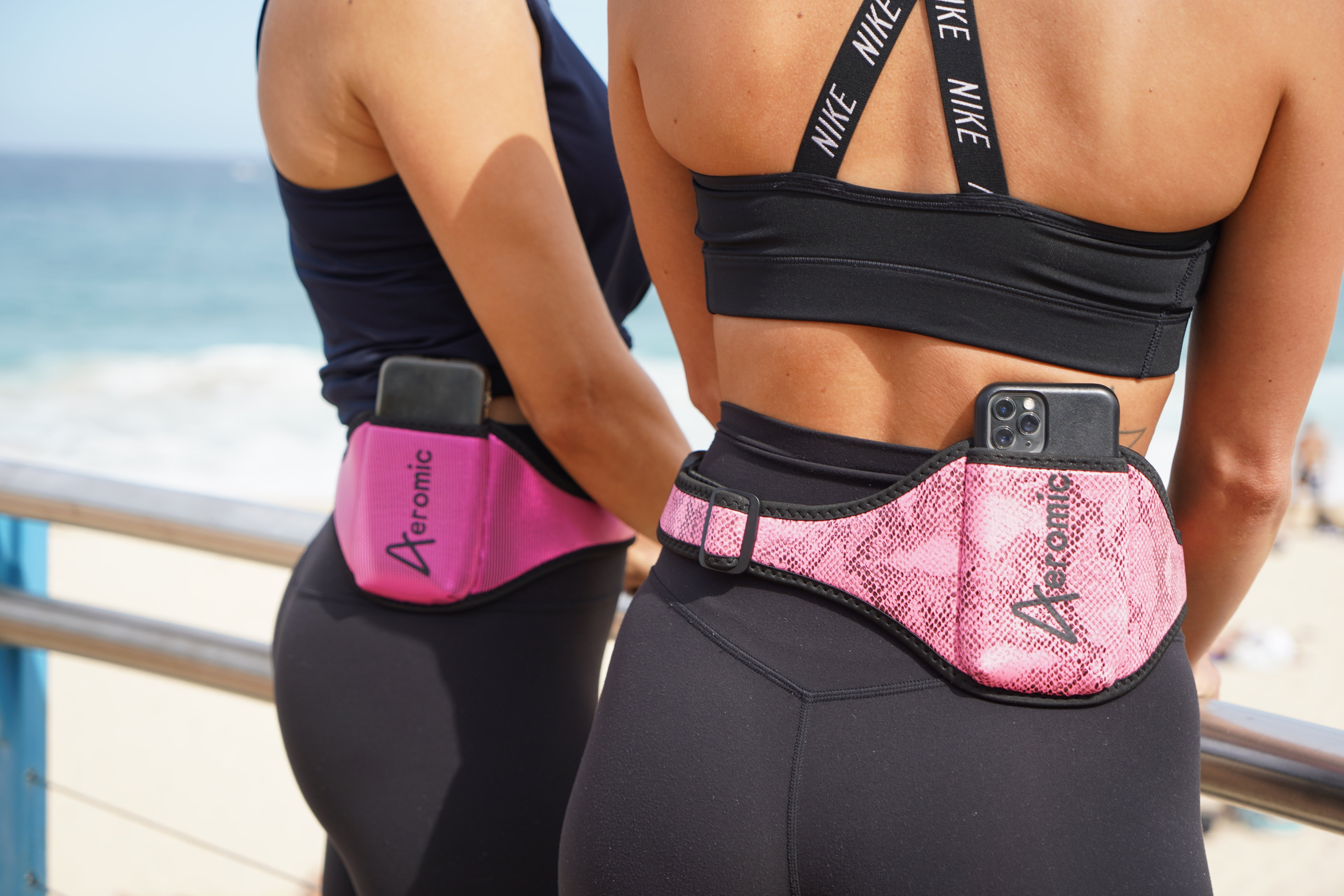 Two women standing by the ocean outdoors, wearing athletic wear, and two pink Aeromic pouch belts on each of their waists.