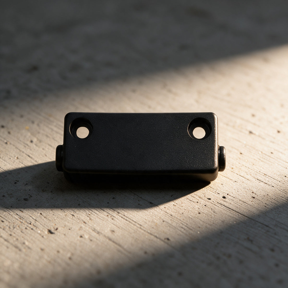 Close-up of the black MTU8 replacement mounting bracket with its two countersunk screw holes and a pivot pin at each end, on grey concrete