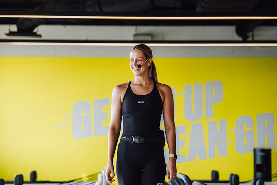 Woman in athletic wear standing in yellow gym with Pilates reformers behind her. Pictured with a yellow Aeromic on her head. 