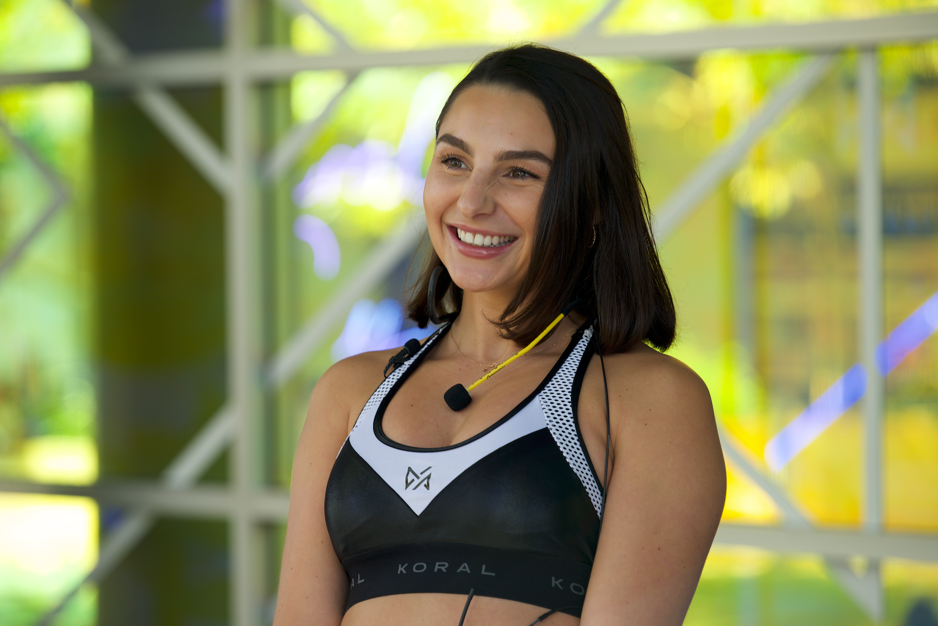 Woman wearing a sports bra with a visible brand logo, standing in front of a colorful geometric background. Pictured with Aeromic around her neck.
