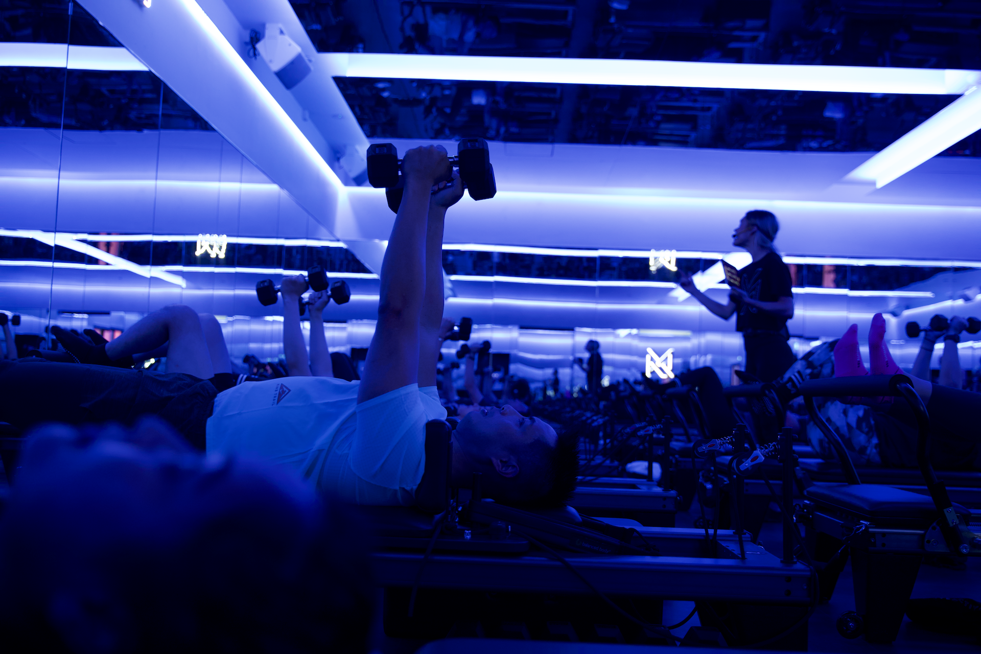 People exercising with dumbbells in a gym with blue lighting. Instructor with headset directing class in background.