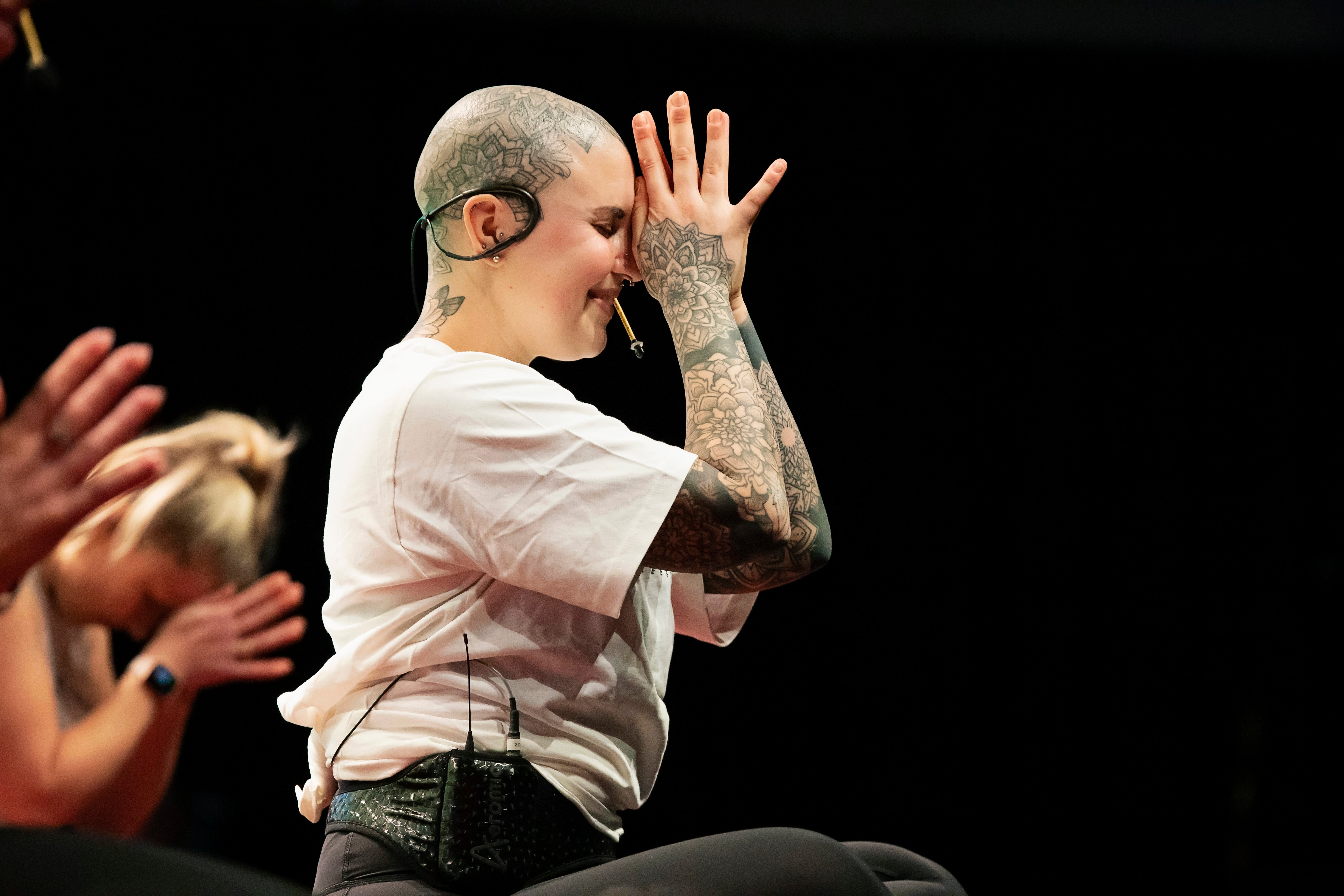 Fitness Instructor with tattoos on stage, wearing a white shirt and black pants, sitting cross legged hands together pressed to face with yellow Aeromic on head.